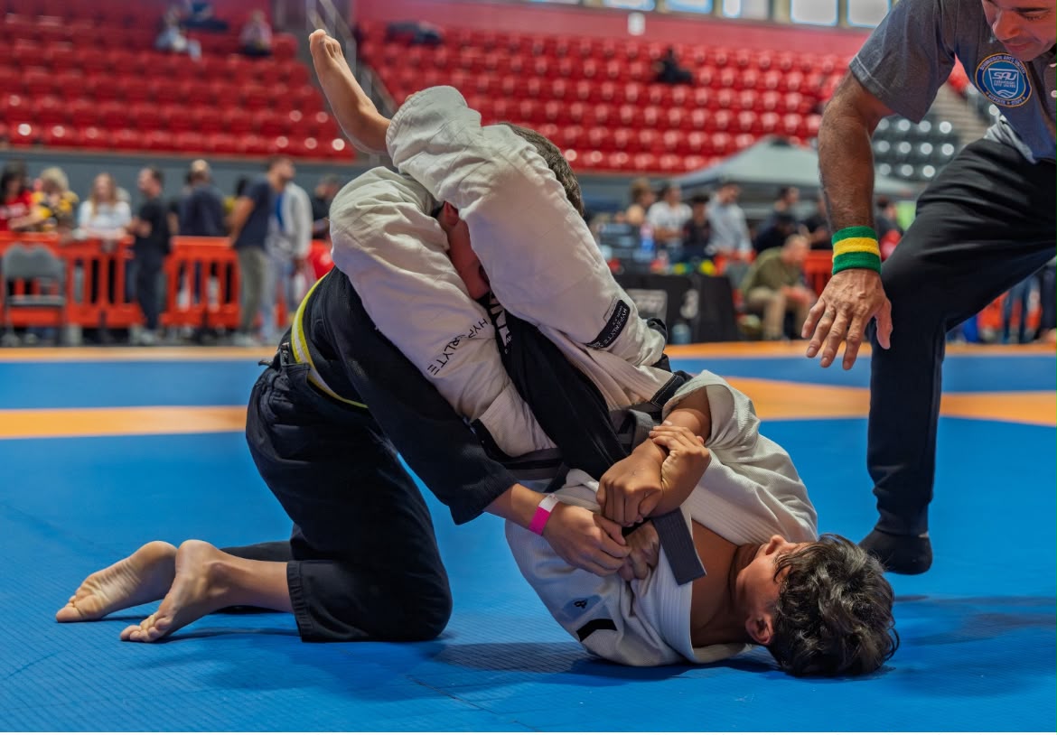 Two youth Jiu Jitsu competitors grappling on a blue competition mat as a referee observes
